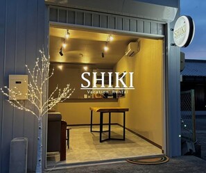 L’entrée d’un hôtel, avec un comptoir de réception, une enseigne indiquant « SHIKI, location de vacances », et un petit arbre décoré de guirlandes lumineuses.