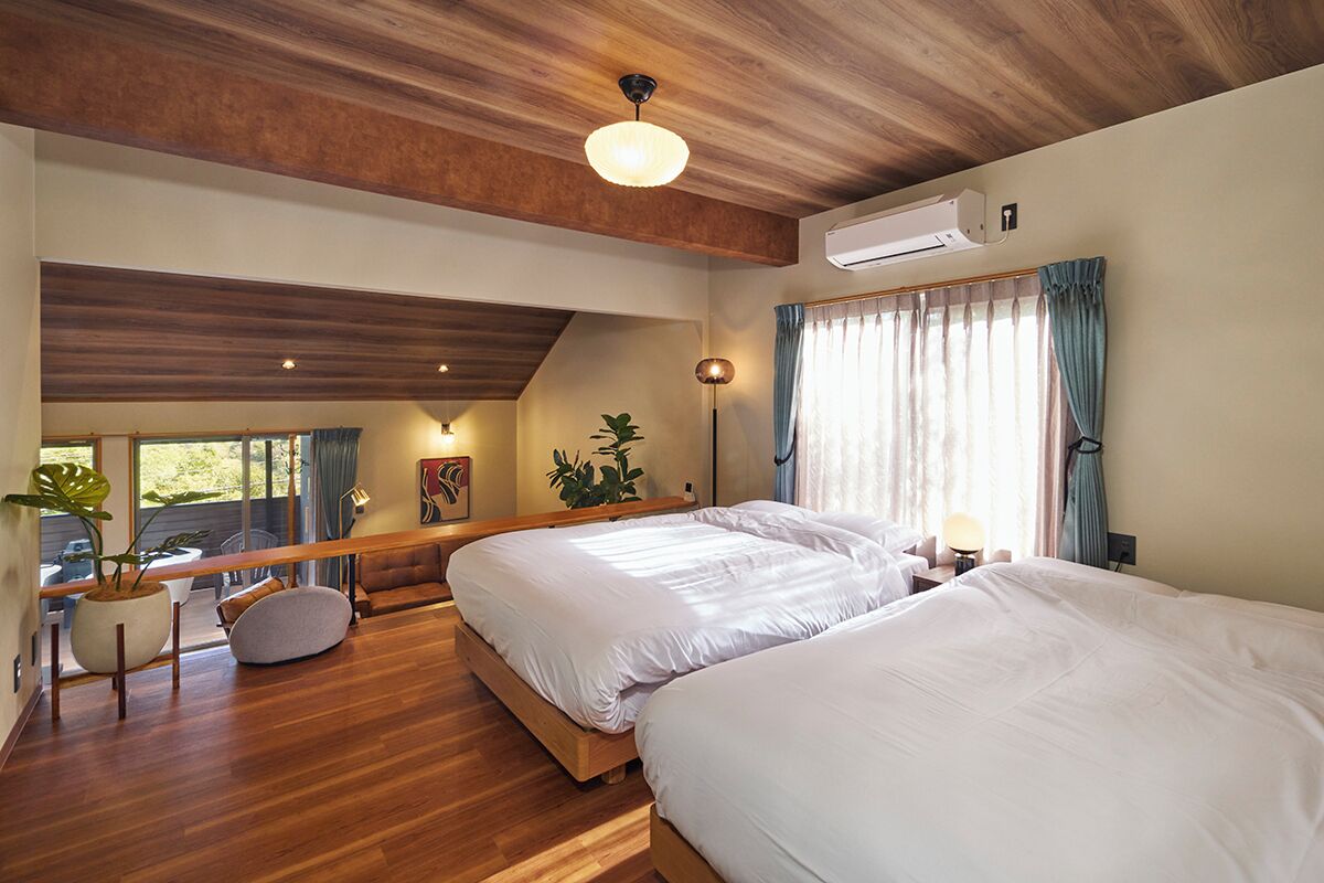 A modern bedroom with two beds, a large window with curtains, a potted plant, and a ceiling fan.