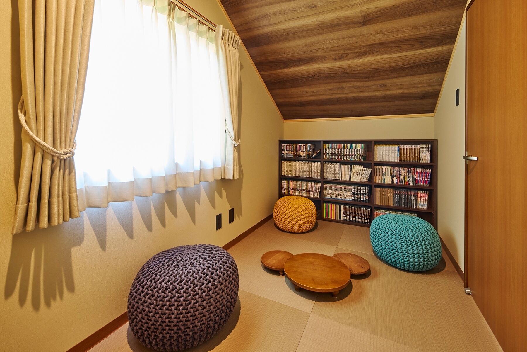 A cozy reading nook with a wooden ceiling, a bookshelf filled with books, and two knitted poufs.