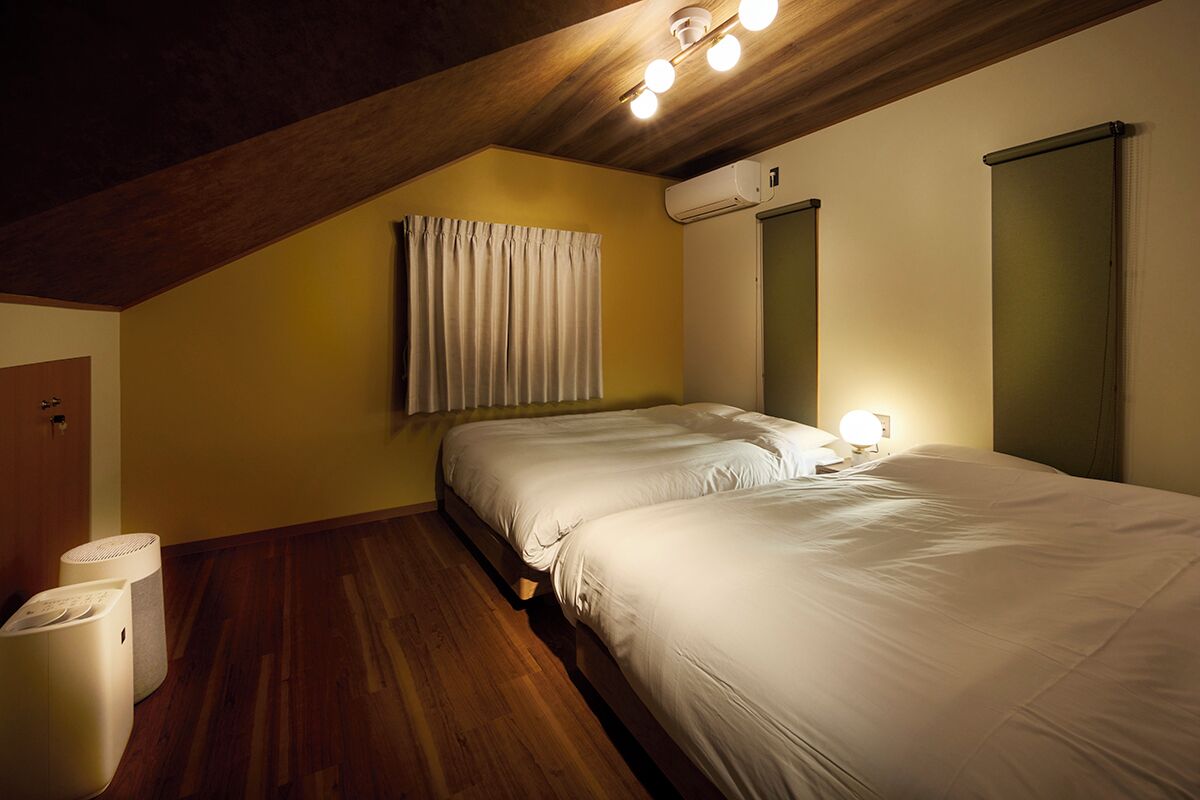 Un dormitorio con techo inclinado, cama con ropa de cama blanca, unidad de aire acondicionado y mesita de noche con lámpara.