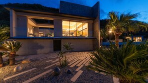 Uma casa moderna de dois andares com amplas janelas de vidro, fachada de pedra e um jardim bem cuidado com palmeiras.