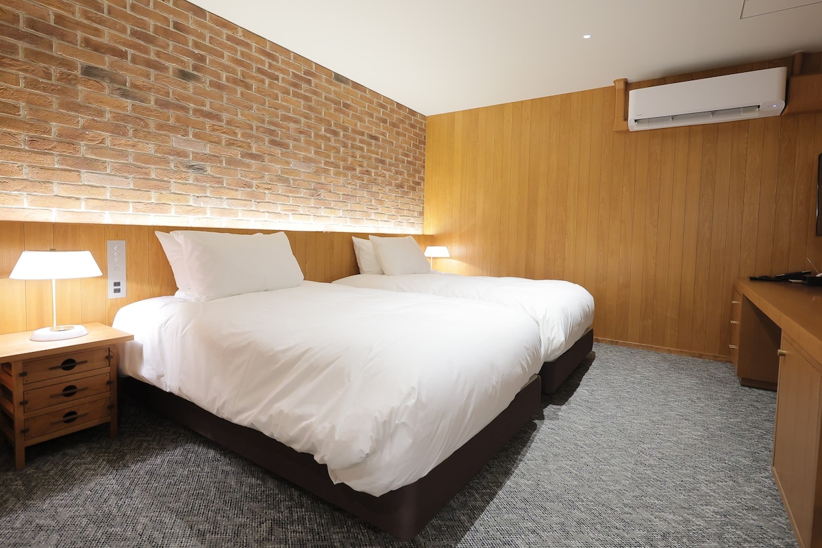 Habitación de hotel con pared de ladrillo, paneles de madera, una cama con ropa blanca, una mesita de noche con una lámpara y un escritorio con una silla.