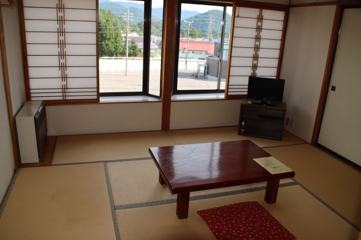 A traditional Japanese room with tatami flooring, a wooden table, a TV on a stand, and a sliding door with a view of the outdoors.