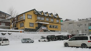 Un immeuble jaune à plusieurs étages, avec un escalier menant à l’entrée, entouré d’un sol recouvert de neige et de voitures garées.