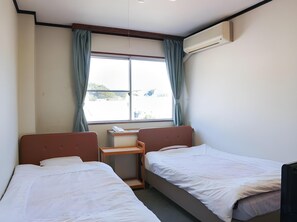 A hotel room with two single beds, a small bedside table, a desk, and a window with curtains. - Maruya Hotel (Tsushima)