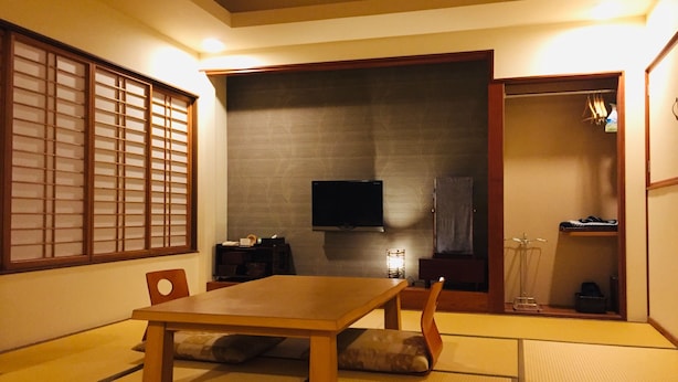 A traditional Japanese room with tatami flooring, a low table, and a television mounted on the wall. - Marufuku Ryokan (Yokote)