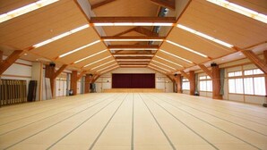 Ein geräumiger Saal mit hoher Decke und Holzbalken, großen Fenstern und einer Bühne im Vordergrund.