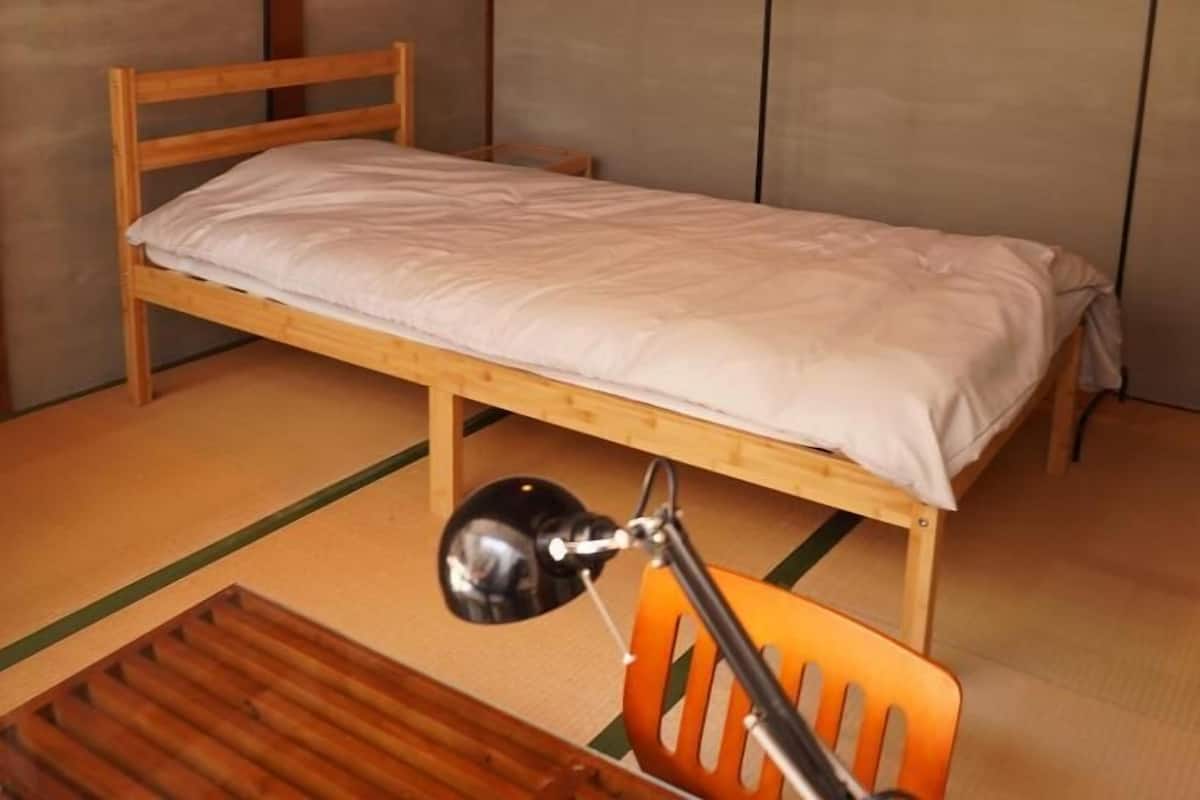 A small, single-bed room with a wooden frame, a white mattress, and a wooden chair with a black armrest.