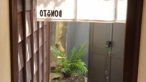 Un étroit couloir extérieur, avec une clôture en bois d’un côté et un portail en métal de l’autre. Au-dessus du portail, on peut lire une enseigne indiquant « DOMOTTO ». Le sol est recouvert de petits cailloux, et une petite plante se trouve près du portail.