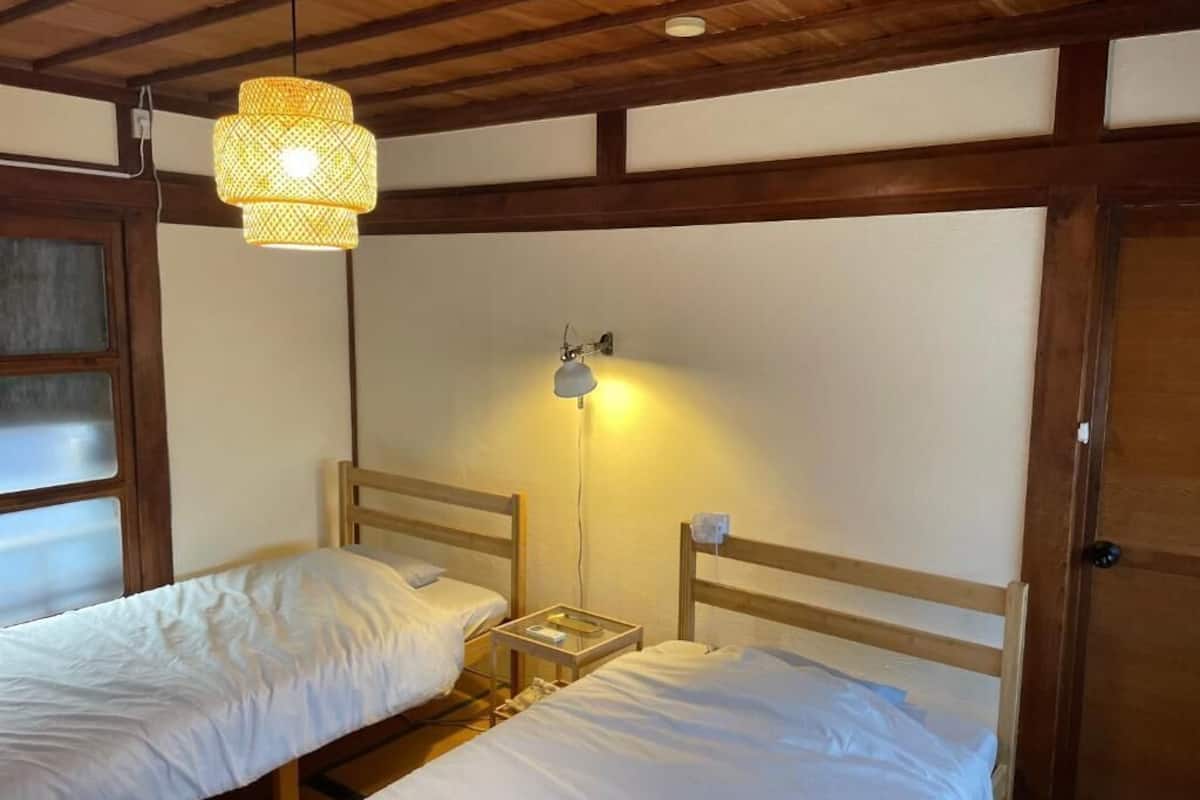 A room with two single beds, wooden headboards, a hanging light fixture, a wall-mounted lamp, and a wooden door.
