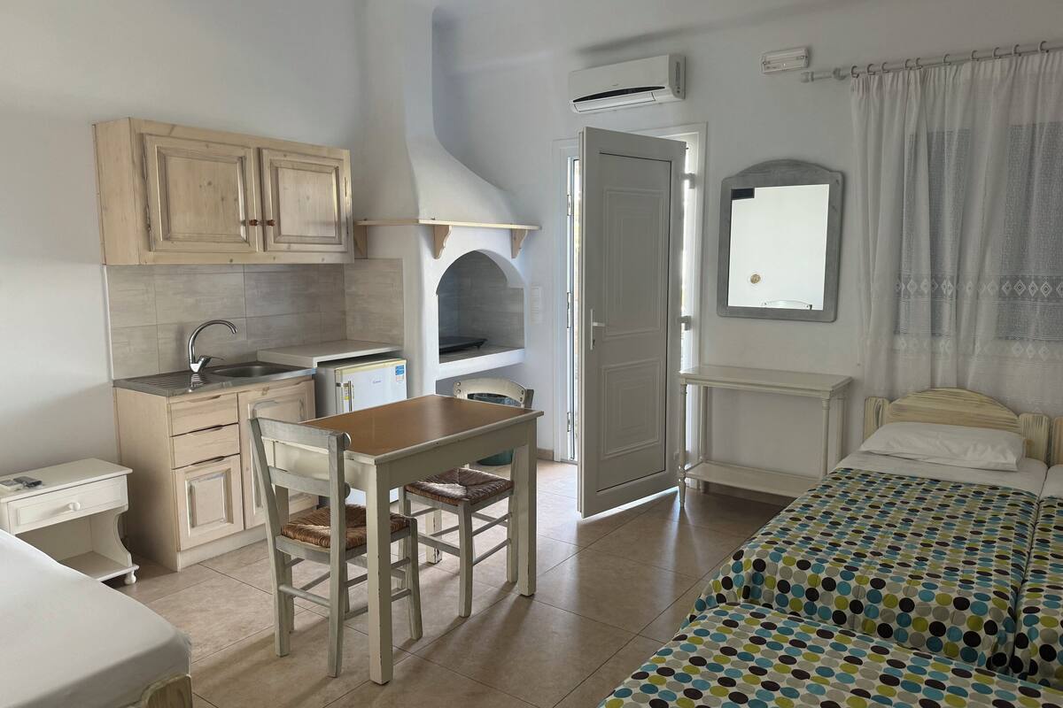 A compact room with a kitchenette, a dining area with a table and chairs, a bed with a patterned bedspread, and a window with curtains.