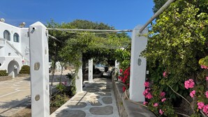 A stone pathway leading to a white building with arches and columns, surrounded by greenery and flowering plants. - Anais Anais Mykonos (Mykonos)