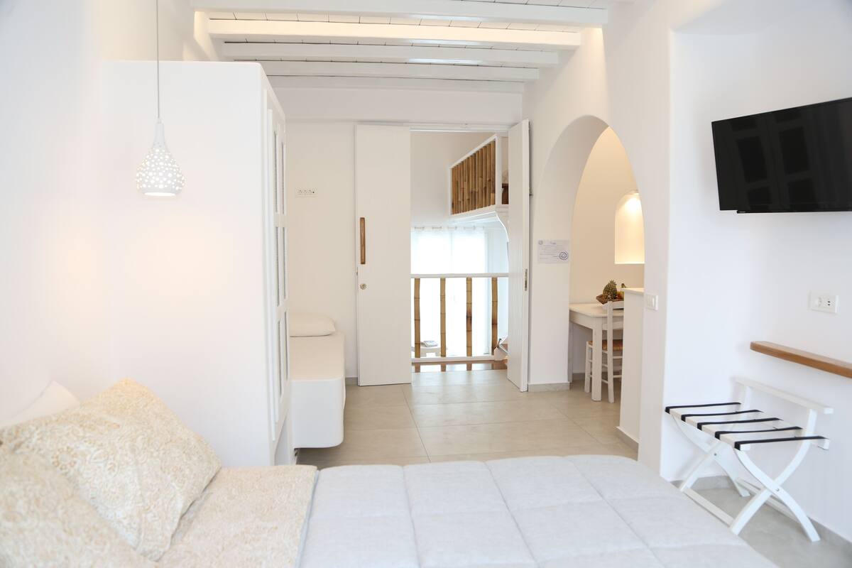 A modern, well-lit room with a white sofa, a hanging light fixture, a TV mounted on the wall, and a view into the kitchen area.
