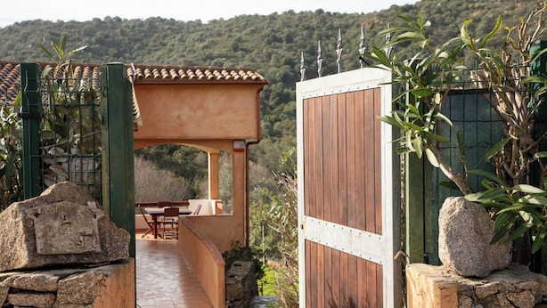 A wooden gate leading to a covered outdoor seating area with a stone wall and a view of a forested hillside. - Casa Leo by Wonderful Italy (Osseddu)