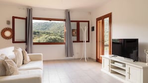 A living room with a sofa, a TV, a stand, and a view of a green landscape through large windows. - Casa Leo by Wonderful Italy (Osseddu)
