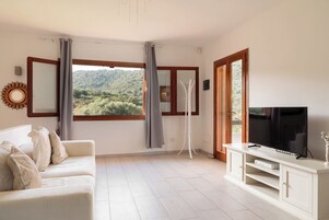 Una sala de estar con sofá, televisor, mueble bajo y vista a un paisaje verde a través de amplias ventanas.