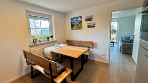 Een eethoek met een houten tafel en banken, een raam met uitzicht en een zichtbare keuken door een open deur.