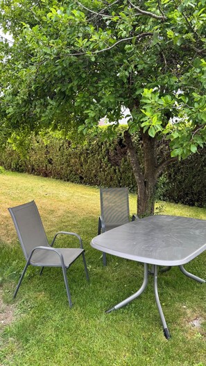 A metal table and two chairs in a grassy outdoor area with a tree and hedge in the background.