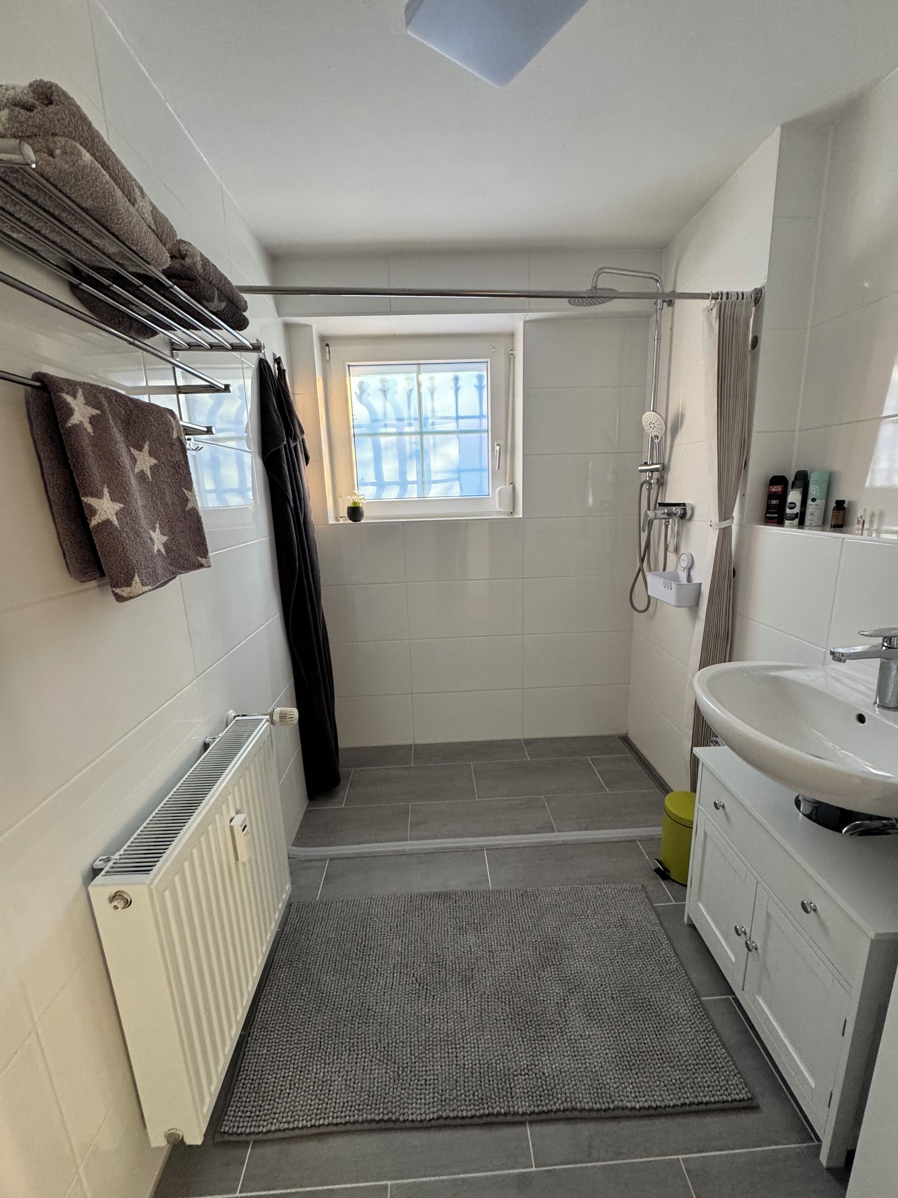 Ein kleines Badezimmer mit einem weißen Waschbecken, einer Dusche, einem Heizkörper und einem kleinen Fenster.