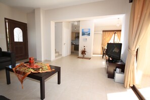 A living room with a dark wooden coffee table, a sofa, and a television. There is a kitchen in the background with cabinets and a countertop. - Luxury Villa Rental, Two Bedroom Villa With Private Pool and Garden, (Pissouri)