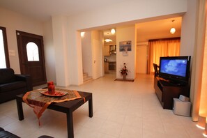 A living room with a dark brown sofa, a coffee table with a decorative centerpiece, a television on a stand, and a kitchen visible through an open doorway. - Luxury Villa Rental, Two Bedroom Villa With Private Pool and Garden, (Pissouri)