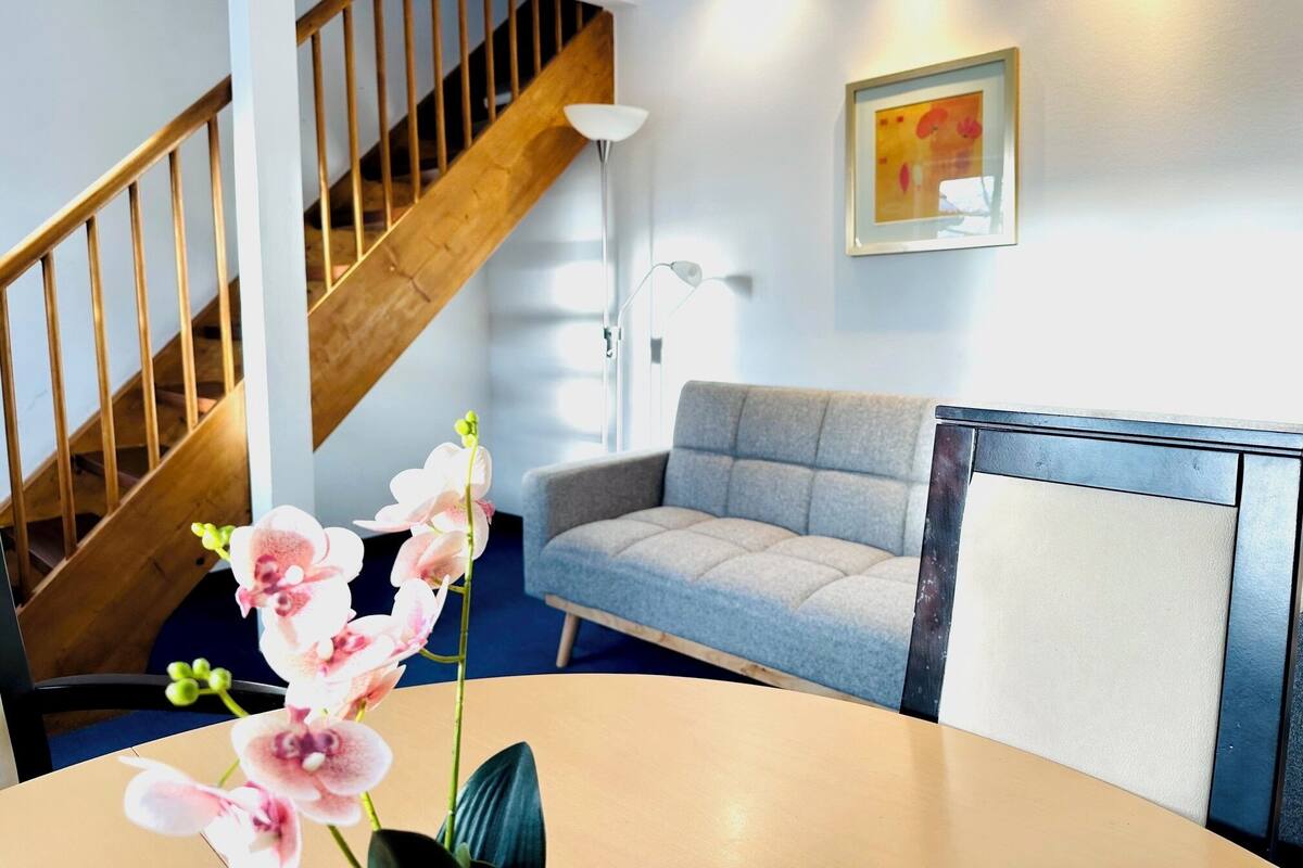 A room with a wooden staircase, a grey sofa, a round table with orchids, and a framed picture on the wall.