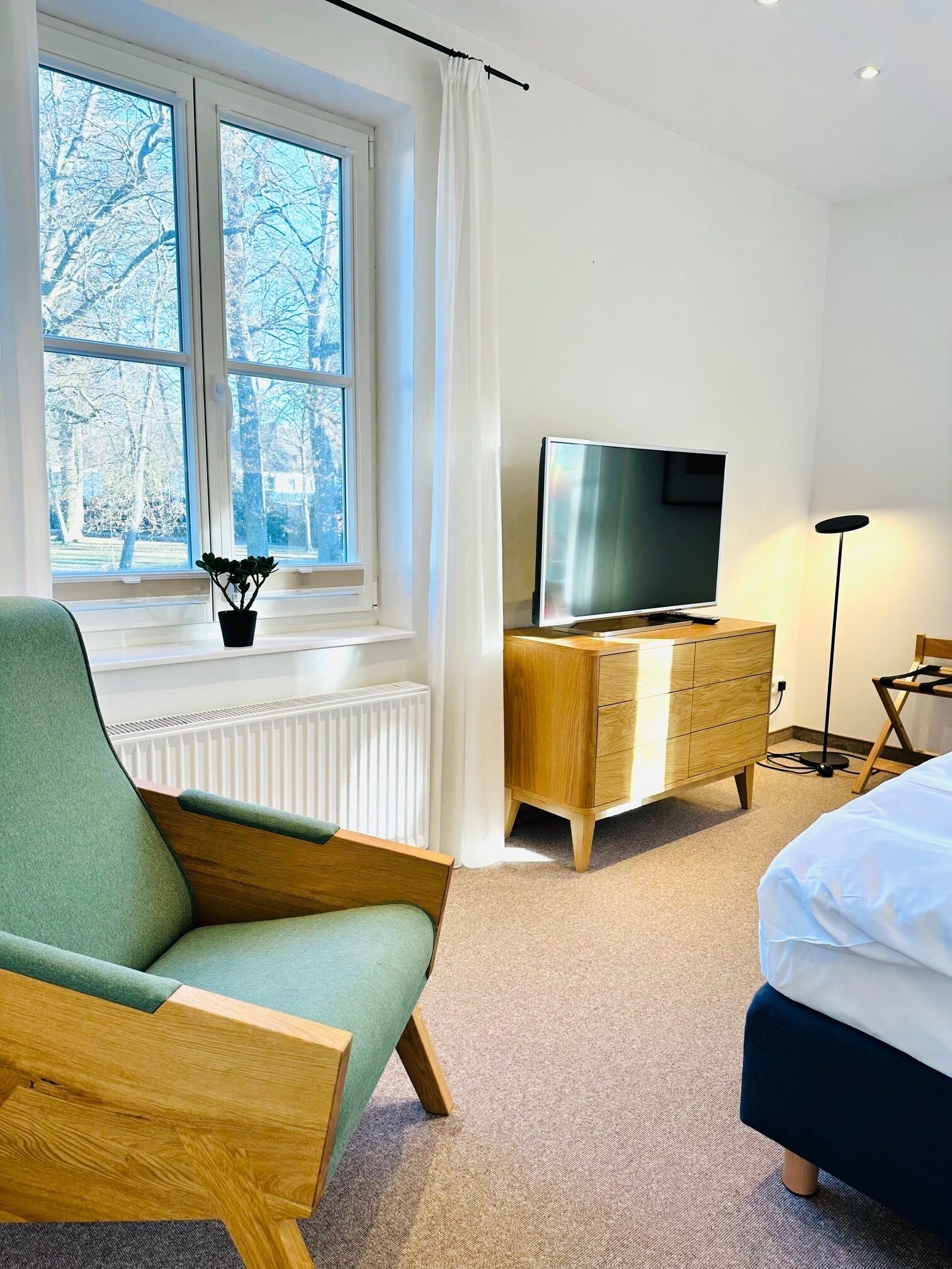 A bedroom with a bed, a TV, a wooden dresser, a chair, a radiator, and a window with a view of trees.