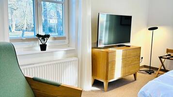 A bedroom with a bed, a TV, a wooden dresser, a chair, a radiator, and a window with a view of trees.