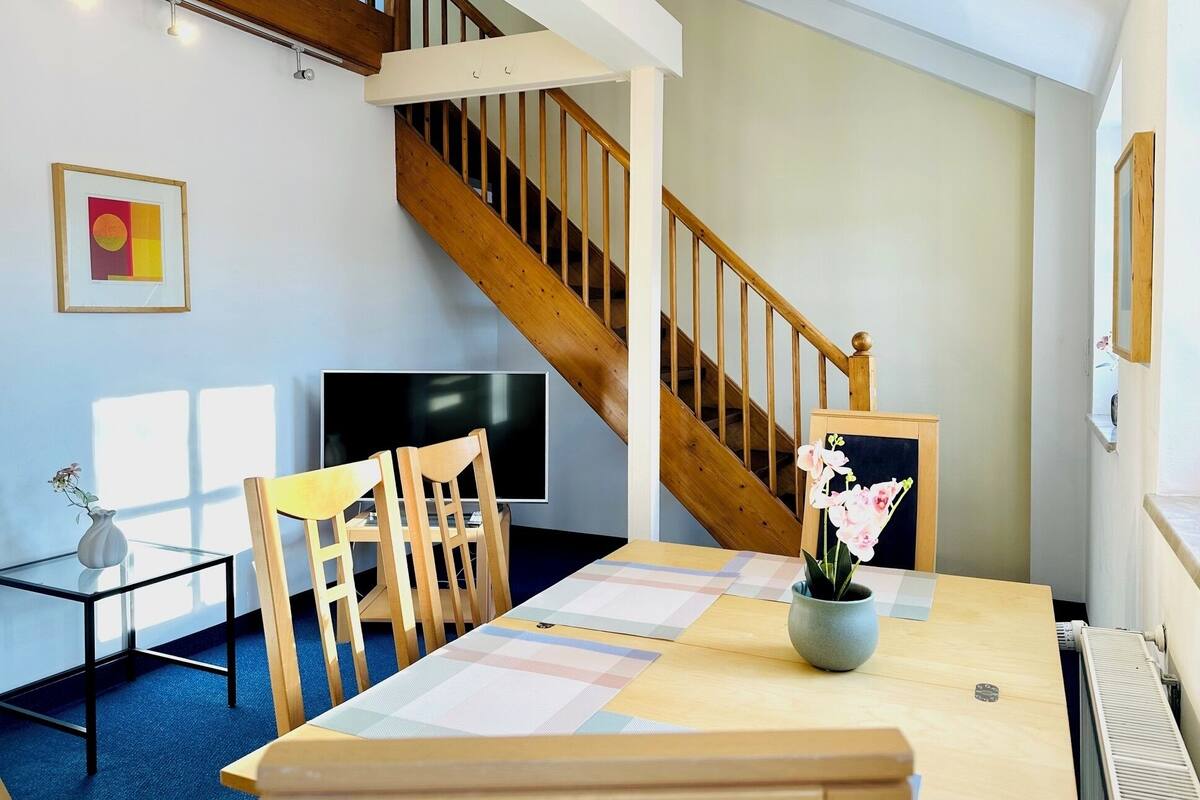 A modern interior with a wooden staircase, a dining area with a table and chairs, a flat-screen TV, and a framed picture on the wall.