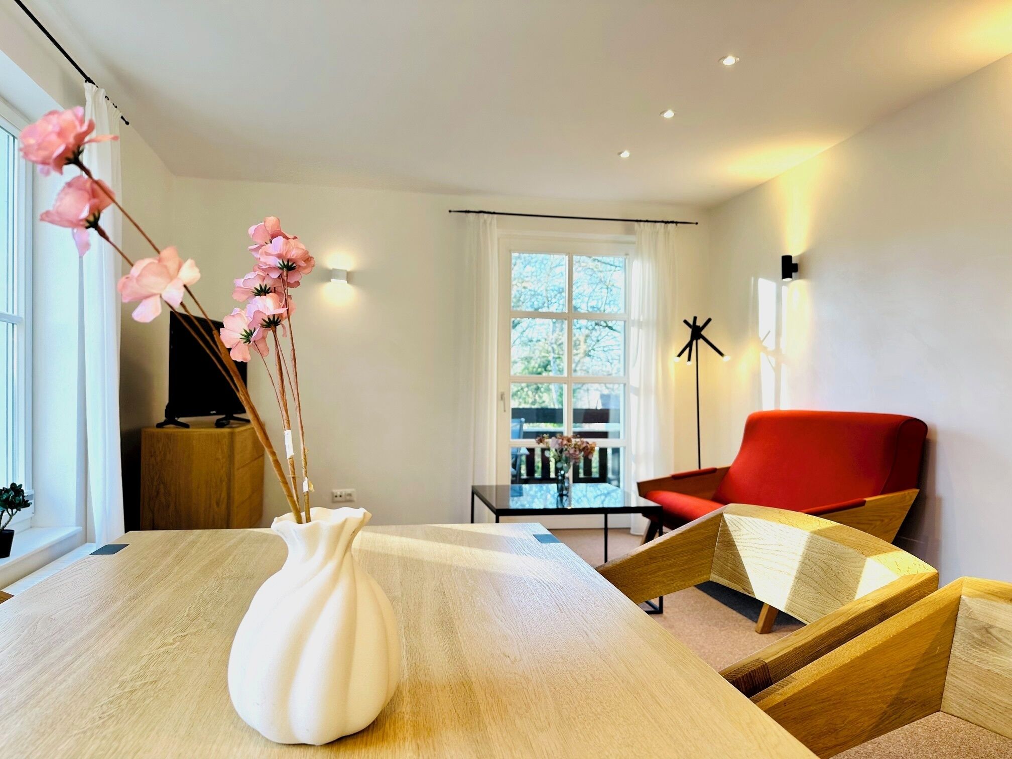 Een moderne woonkamer met een houten eettafel, een witte vaas met roze bloemen, een rode stoel en uitzicht op het groen buiten.