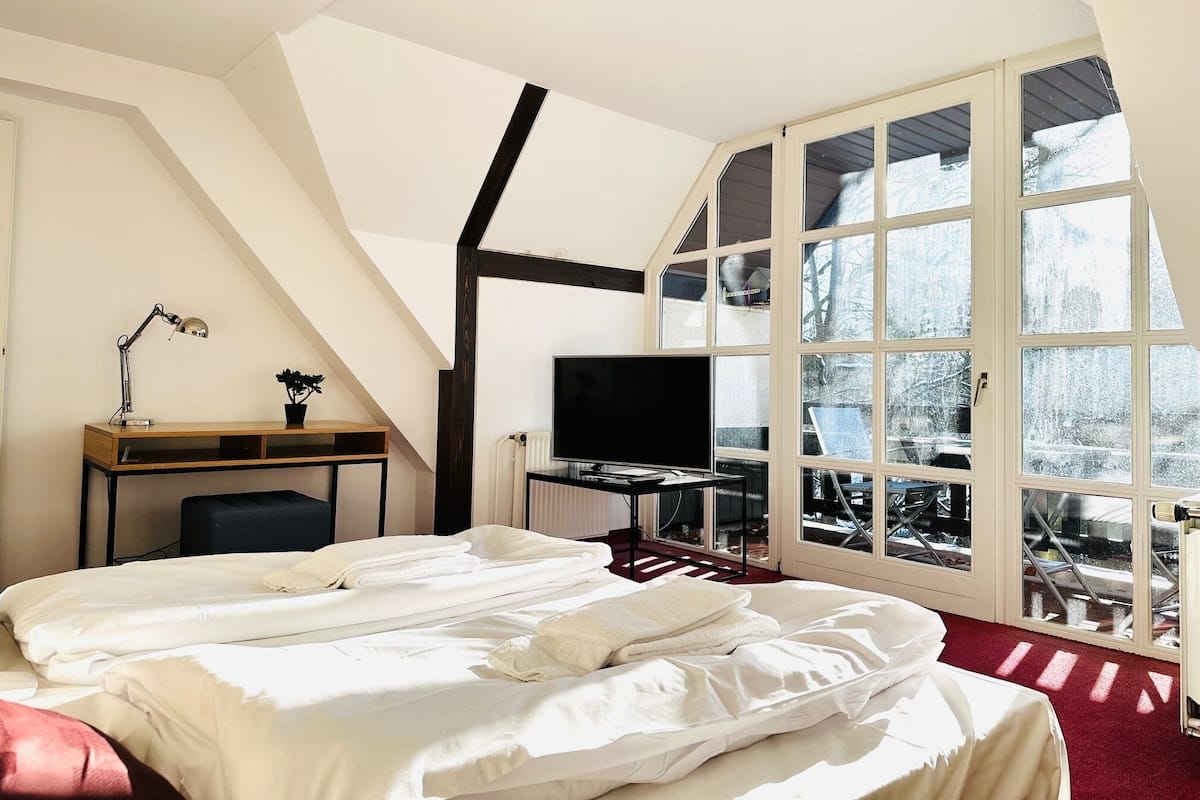 A bedroom with a bed, a desk with a computer, a TV, and large windows.