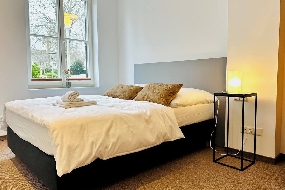 A neatly made bed with a white duvet and two patterned pillows, a bedside table with a lamp, and a window with a view of trees and buildings.