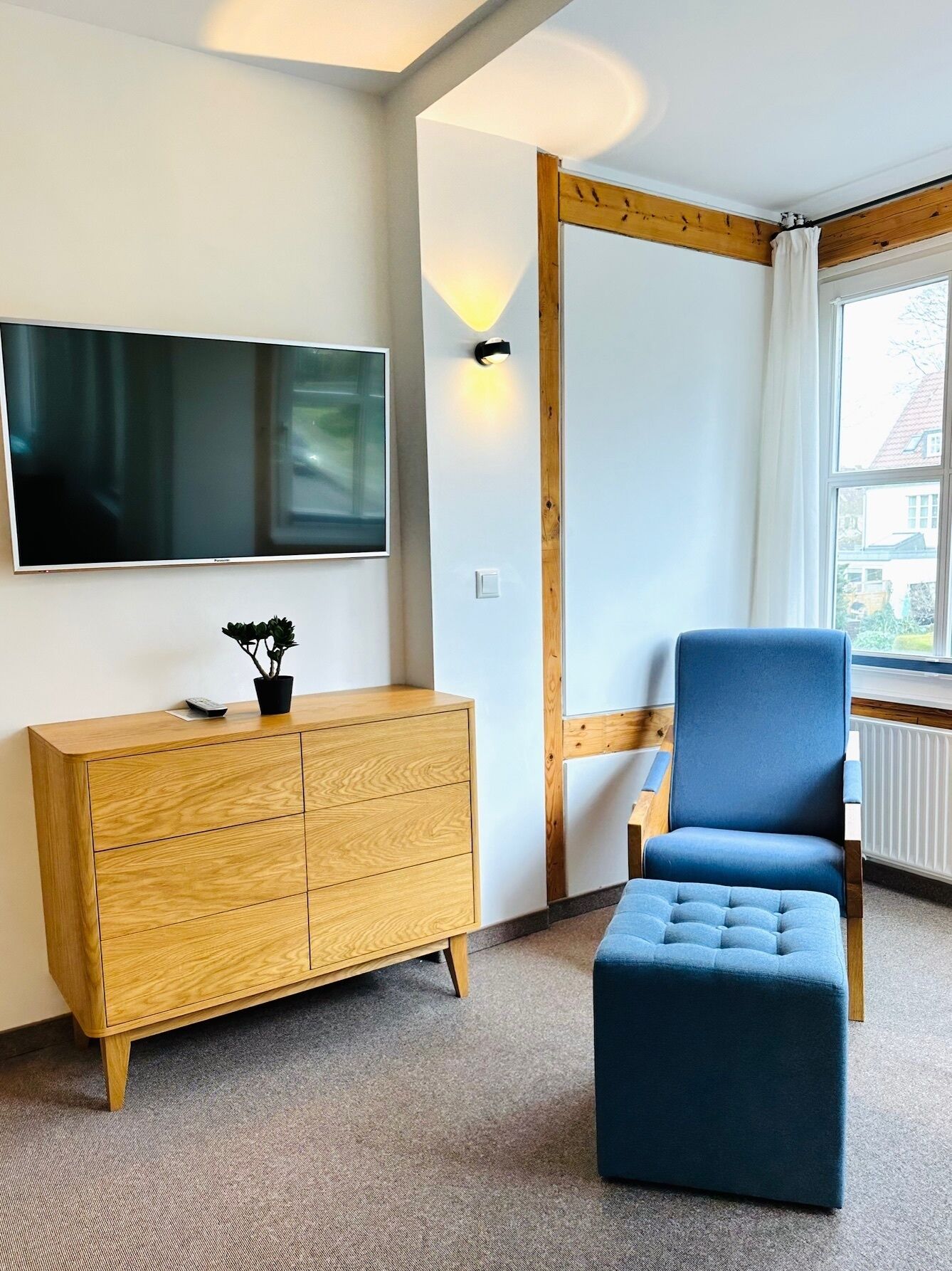 A room with a wooden cabinet, a blue chair, a television mounted on the wall, and a window with curtains.