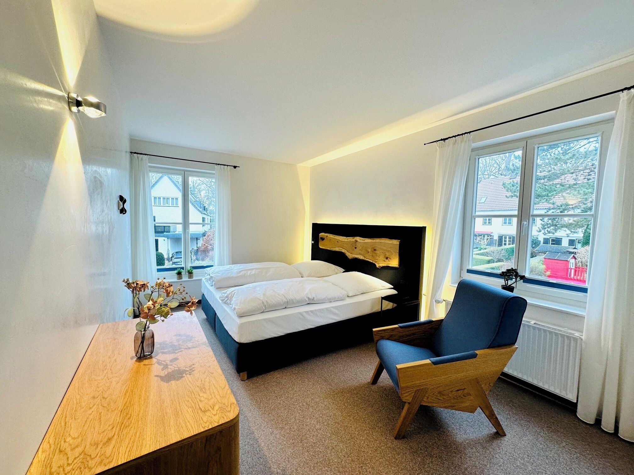 A bedroom with a bed, a chair, a desk, and a window with a view of houses.