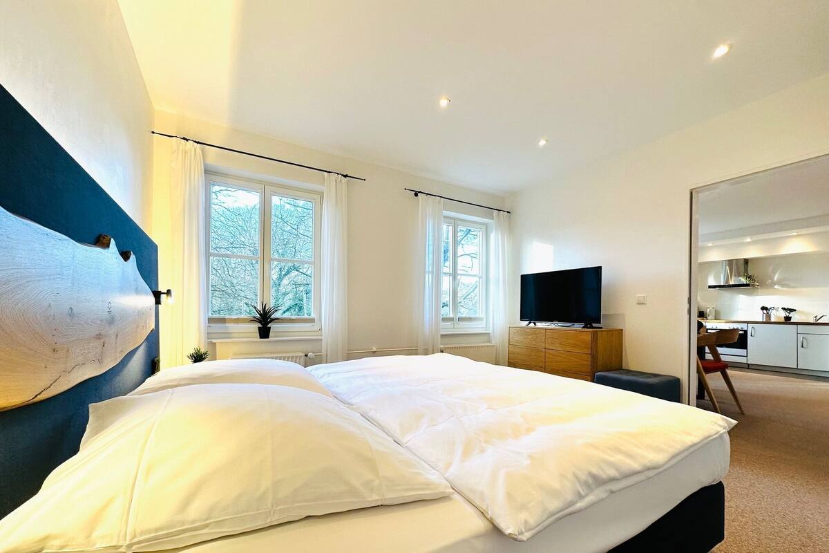 A modern bedroom with a large bed, a flat-screen TV, and a view of the outdoors through the windows.