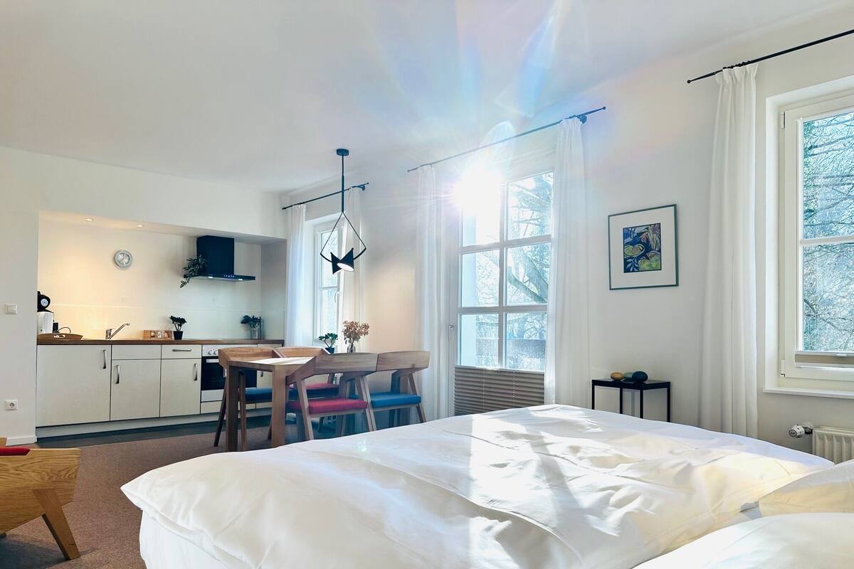 A modern bedroom with a large bed, a dining area with a table and chairs, a kitchen in the background, and a window with a view of trees.