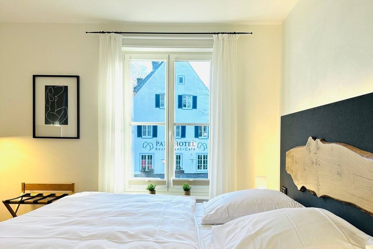 A hotel room with a bed, a window with curtains, a framed picture on the wall, and a view of a building outside.