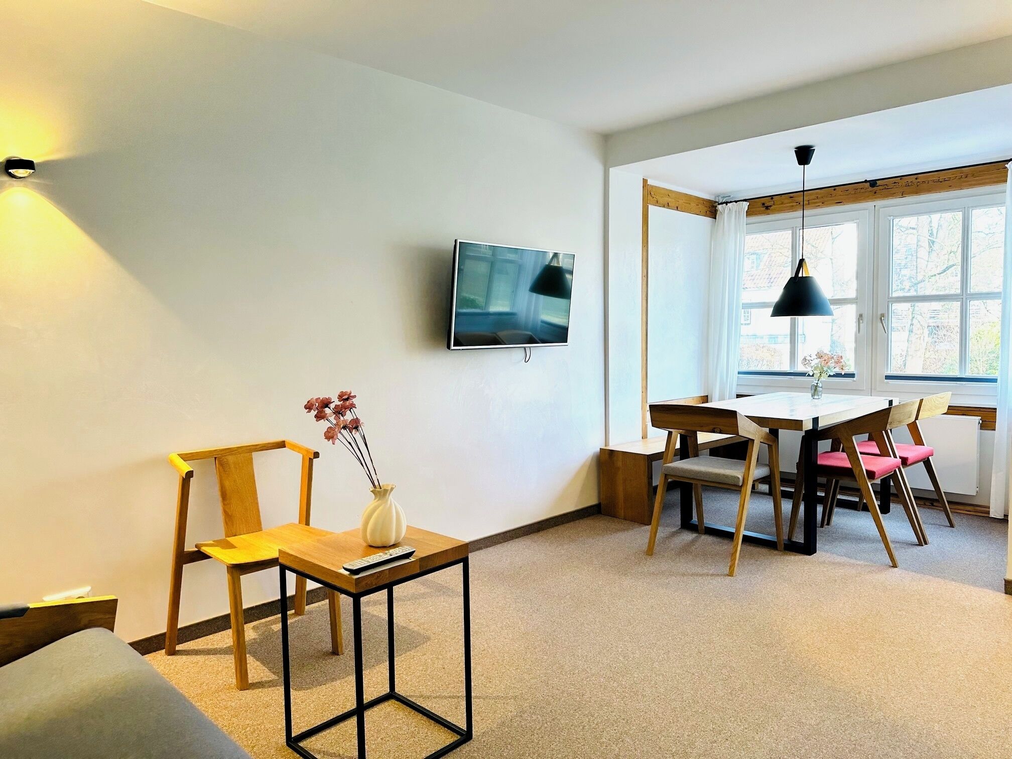 A modern living room with a TV, a dining table, chairs, a small side table with a vase, and a window with curtains.