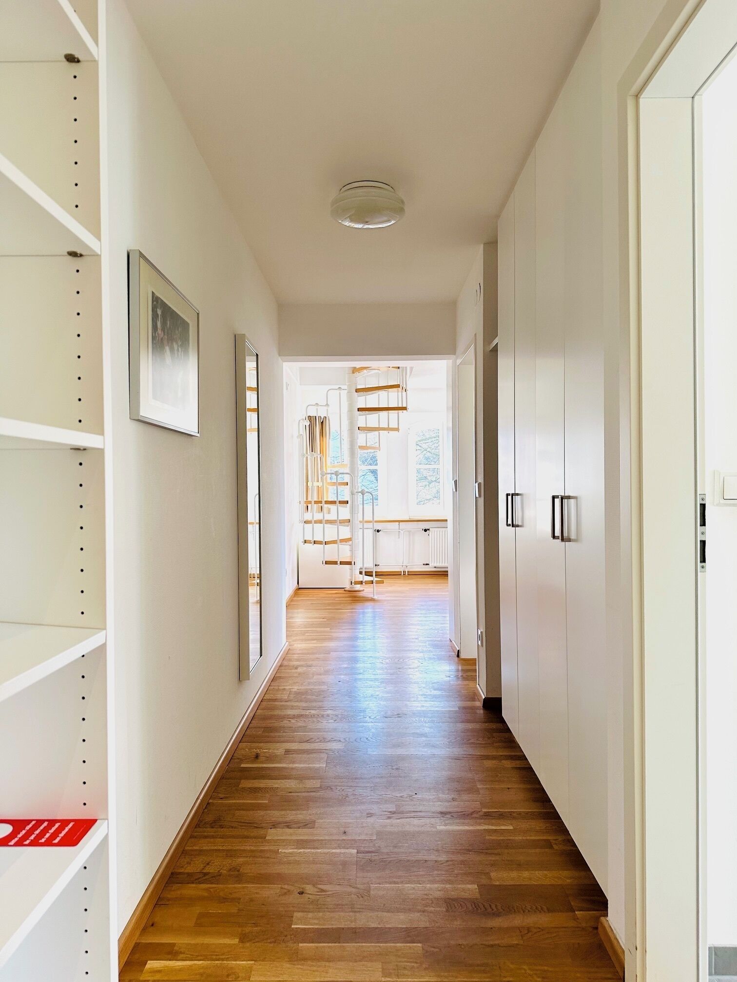 A hallway with wooden flooring, white walls, and a staircase leading to a bright room with windows.