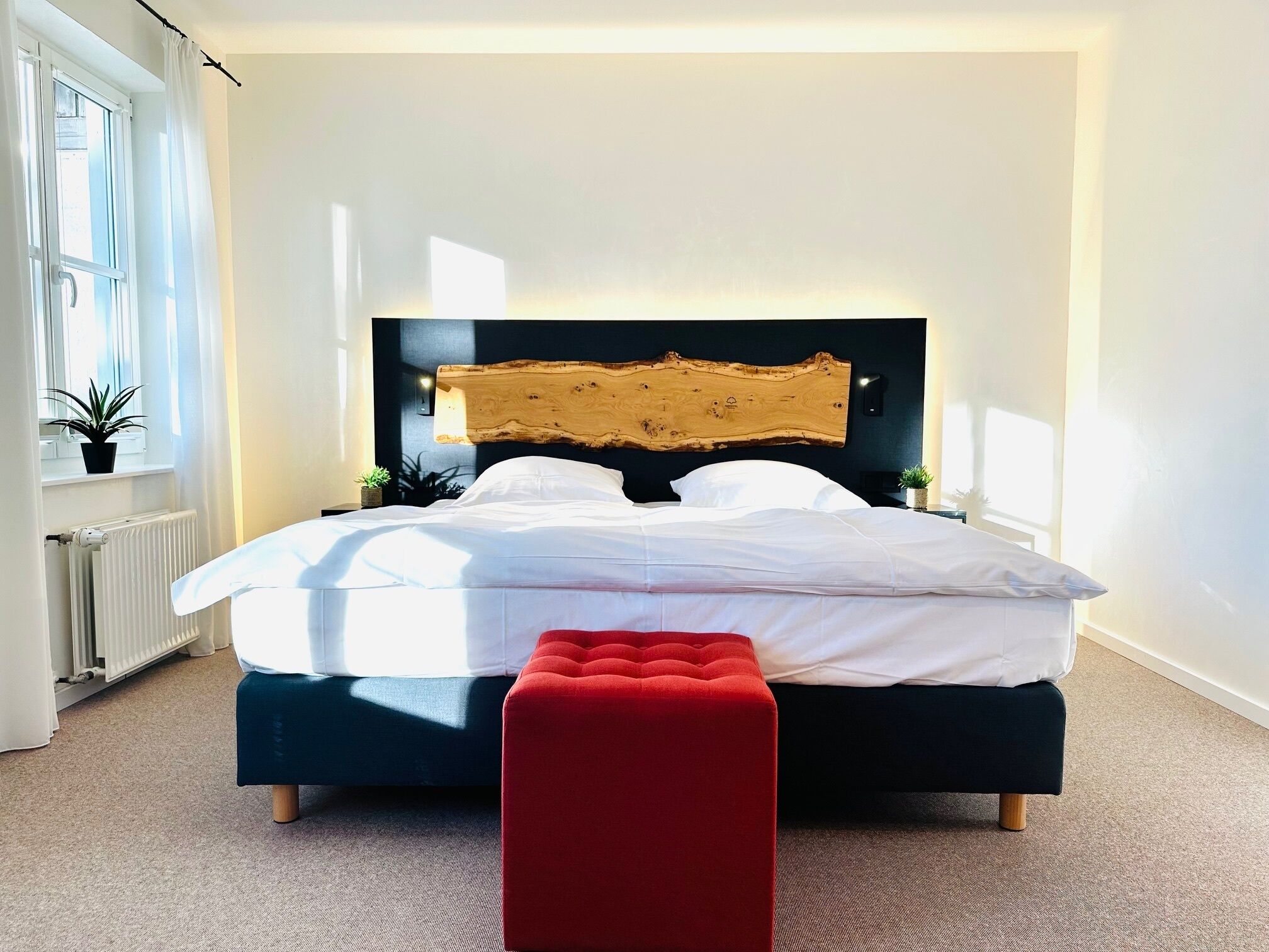 A modern bedroom with a large bed, a red ottoman, and a wooden headboard.
