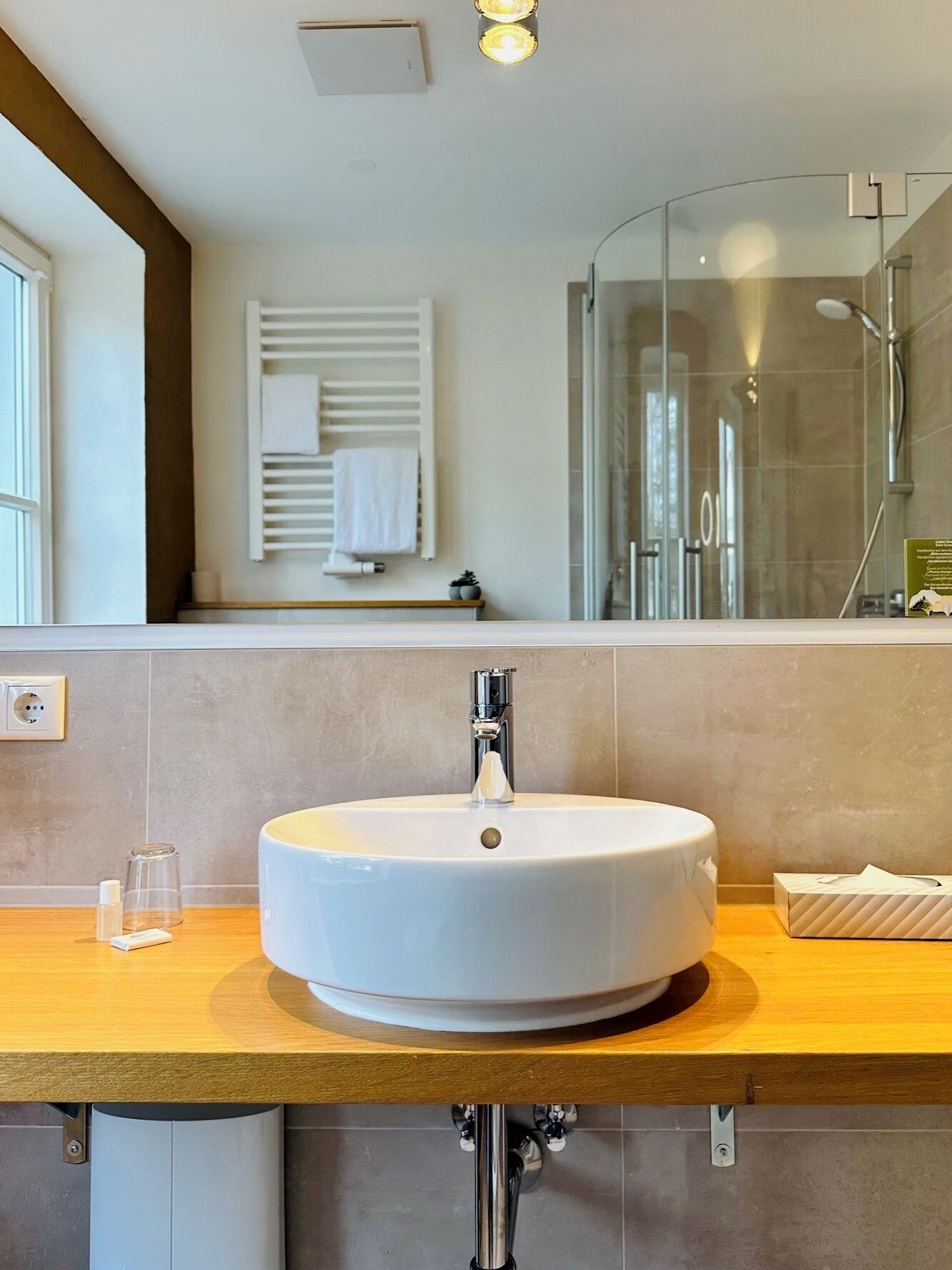 Un baño moderno con un lavabo redondo, una cabina de ducha con paredes de vidrio y un perchero con una toalla blanca.