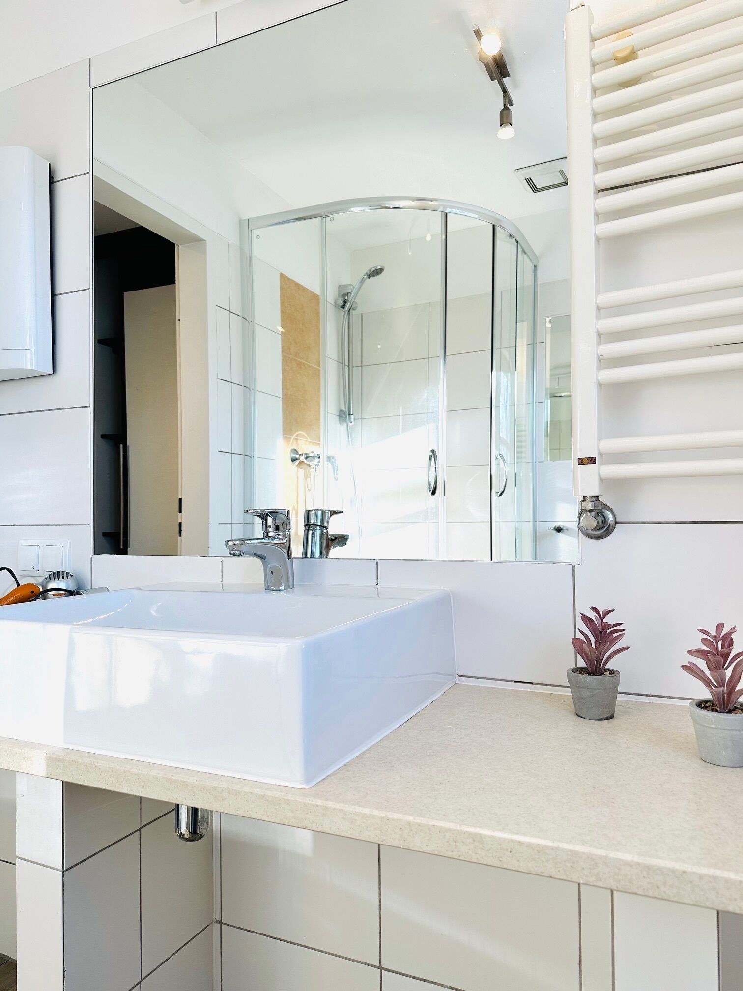 A modern bathroom with a large mirror, a shower enclosure, a sink, and two potted plants.