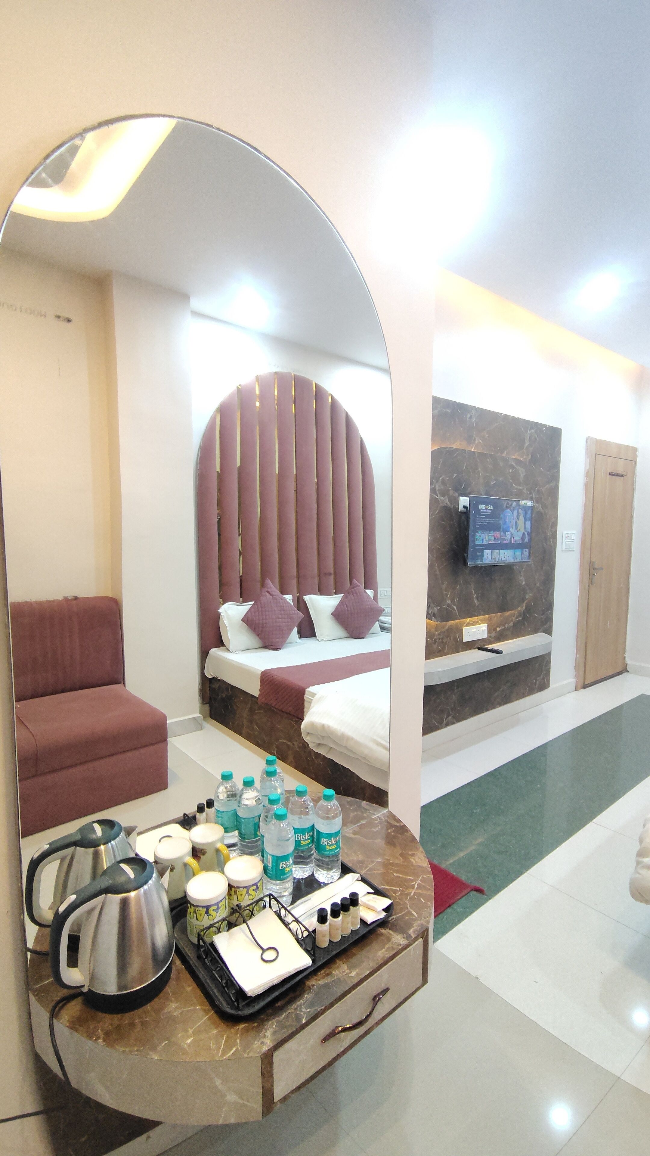 A hotel room with a bed, a sitting area, a mirror, a TV, and a tray with toiletries.