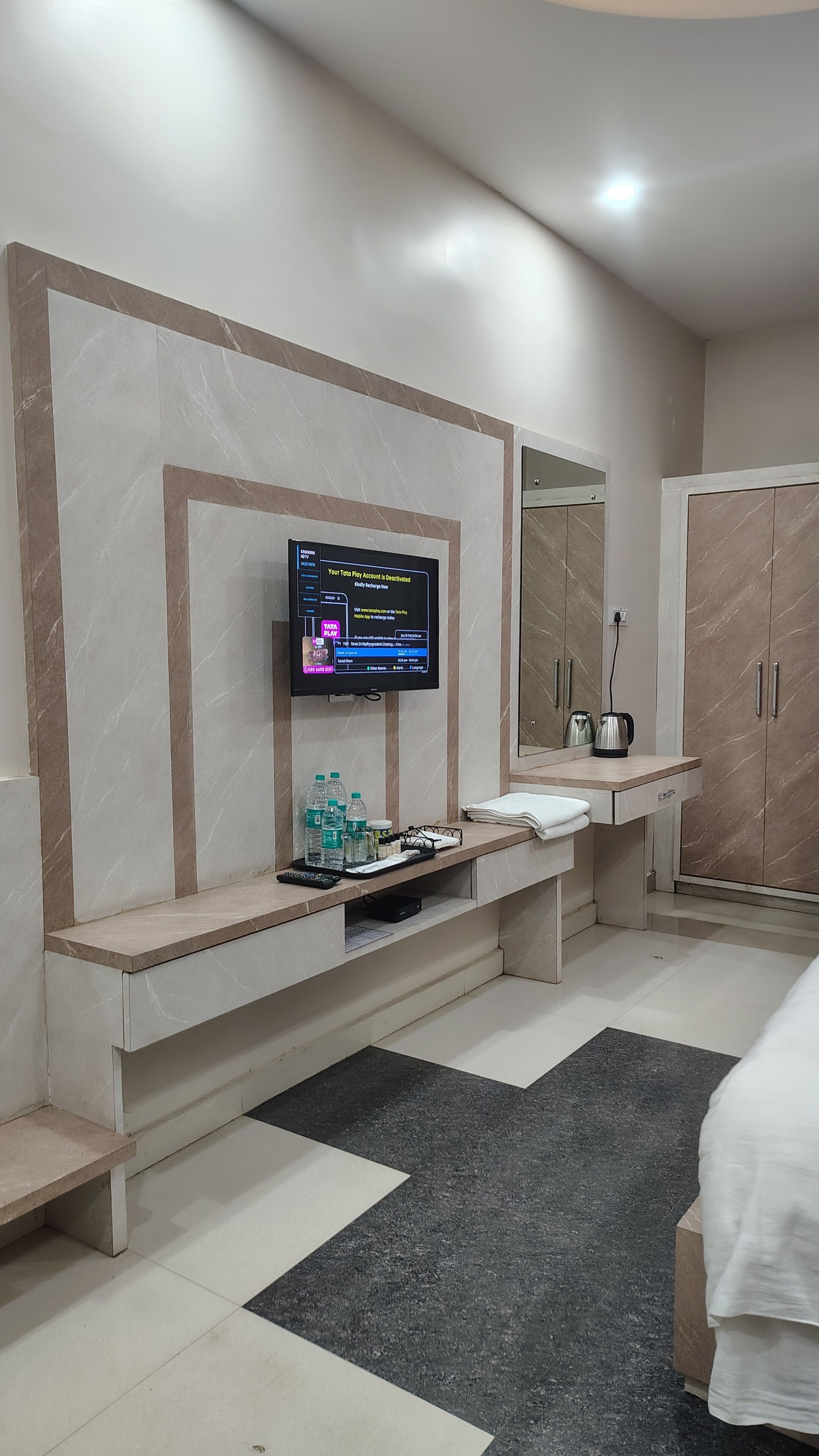 Una habitación de hotel moderna con un televisor de pantalla plana montado en la pared, un tocador con espejo, un banco de madera y un armario.