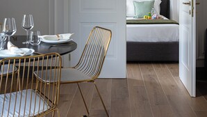 Ein modernes Zimmer mit Essbereich, ausgestattet mit einem runden Tisch, goldenen Stühlen und einer weißen Tür zu einem Schlafzimmer mit Bett, Kissen und Nachttisch.