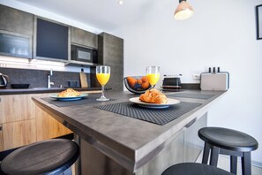 Eine moderne Küche mit einer zentralen Kochinsel, zwei Barhocker und einer Frühstücksvorrichtung mit Croissants und Orangensaft.