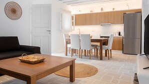 Un salon moderne avec un canapé, une table basse en bois et un coin repas comprenant des chaises et une table. On aperçoit à l’arrière-plan une cuisine avec des meubles en bois et un réfrigérateur.