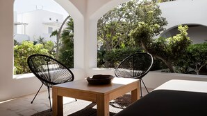 A patio with two black chairs, a wooden table, and a bowl. It features white walls and arches, with a view of greenery and a building in the background. - Mykonos pool house 5 shared pool - My rental Homes (Mykonos)