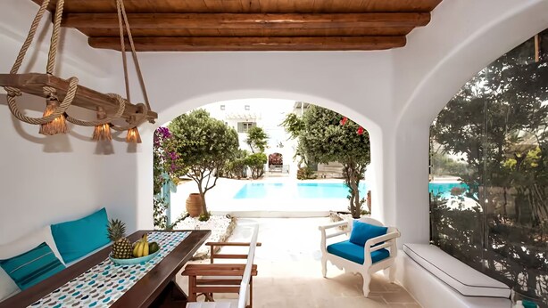 Un patio iluminado por el sol con vista a la piscina, una mesa de comedor con piña y plátanos, y un sofá blanco con cojines azules.