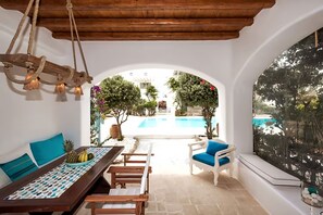 A sunlit patio with a pool view, a dining table with a pineapple and bananas, and a white sofa with blue cushions. - Mykonos pool house 6 shared pool - My rental Homes (Mykonos)